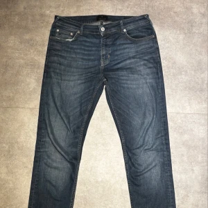 Mörkblå Jens junk de luxe - Snygga mörkblå jeans med klassisk femficksdesign och Flex Fit-passform. Jeansen har slitningar vid fickorna och subtila tvättade detaljer som ger en cool look. Perfekta för dig som gillar en avslappnad stil med lite edge. Storleken är 33/32