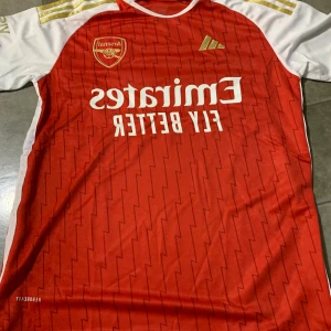 Arsenal röd fotbollströja Adidas S - Säljer en officiell Arsenal fotbollströja från Adidas i storlek S. Tröjan är röd med vita ärmar och har guldiga detaljer på axlarna. Den har ett blixtmönster över hela framsidan och baksidan, samt Arsenal-logga och Adidas-märke på bröstet. Materialet är lätt och andas.