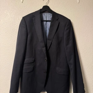 Randig mörkblå kavaj från Morris - Snygg mörkblå kavaj från Morris med diskreta ränder. Klassisk modell med två knappar, tre fickor framtill och elegant ljusblått innerfoder. Tillverkad i 100% ull för en exklusiv känsla. Perfekt för dig som vill ha en stilren och tidlös look. 