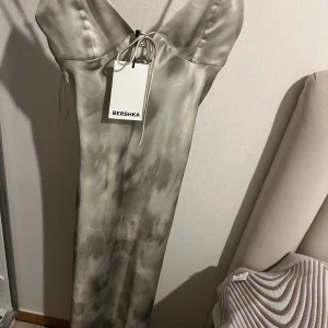 Ljusgrön långklänning från Bershka - Supersnygg långklänning från Bershka i ljusgrön satinlook. Klänningen har smala axelband, v-ringning och en liten knytning framtill. Materialet är glansigt och känns lyxigt, perfekt för dig som vill sticka ut med en elegant vibe.