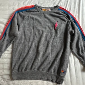Grå sweatshirt Aviator Nation regnbågsränder - Grå sweatshirt från Aviator Nation med färgglada regnbågsränder längs ärmarna och en rosa blixt på bröstet. Tröjan har rund hals, långa ärmar och mjukt material som känns skönt mot huden. Perfekt för dig som gillar retrostil och färg.