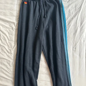 Mörkblå mjukisbyxor Aviator S - Mörkblå mjukisbyxor från Aviator Nation med blå och turkosa ränder längs ena benet. Byxorna har dragsko i midjan och är tillverkade i en mjuk blandning av polyester, bomull och rayon. Perfekta för chill dagar eller när du vill ha en avslappnad look.