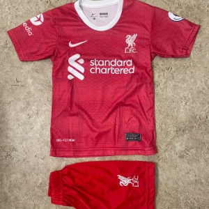 Liverpool Salah #11 fotbollströja Nike - Röd Liverpool fotbollströja med M. Salah och nummer 11 på ryggen. Tröjan har korta ärmar, vit krage och vita tryck med klubbmärke och sponsor. Tillverkad i lätt, ventilerande material med Dri-FIT-teknologi. Matchande röda shorts med LFC-logga ingår.