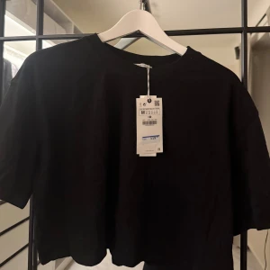 Svart croppad t-shirt från Zara - En svart croppad t-shirt från Zara med rund halsringning och korta ärmar. Enkel och stilren design i mjuk bomull, perfekt att matcha med jeans eller kjol. T-shirten har en avslappnad passform och är ett måste i garderoben.