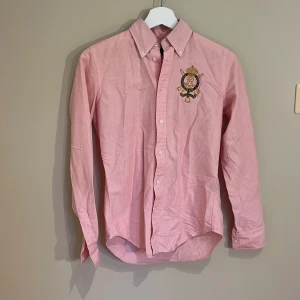 Rosa skjorta från Ralph Lauren - Snygg rosa skjorta från Ralph Lauren med broderad emblem i guld och marinblått på bröstet. Skjortan har klassisk krage, långa ärmar och knappar framtill. Tillverkad i 100% bomull för en skön och fräsch känsla. Perfekt för dig som gillar stilrena detaljer. Storleken 2 motsvarar xs/s🩷knappt använd så inga defekter eller tecken på användning!