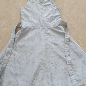 Klänning - Ljusgrå långklänning i glansigt tyg från Lindex Kids, storlek 128. Klänningen har broderade detaljer med rosett och paljetter i midjan samt dragkedja i ryggen. Materialet känns som polyester och modellen har breda axelband och knytband i midjan. Har några små fläckar och bandet bakom har gått av. Annars är den i bra skick.