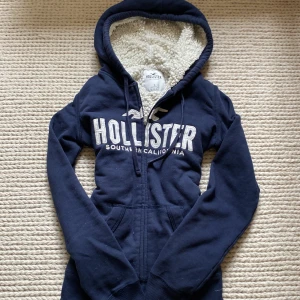 Marinblå hoodie från Hollister S - Pris kan diskuteras!!💗 Mysig marinblå hoodie från Hollister med broderad logga och text på bröstet. Fodrad med fluffigt vitt teddyfoder i huvan, dragkedja framtill och snörning vid huvan. Perfekt för kyliga dagar och riktigt skön att ha på sig. Har ett litet hål vid armhålan men inte något som märks om man inte kollar supertydligt. Lite mindre på bild än i verkligheten 