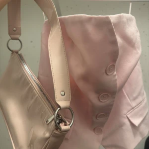 Ljusrosa axelväska med dragkedja och en rosa topp - Snygg ljusrosa axelväska i glansigt syntetmaterial med silvriga metalldetaljer och dragkedja. Perfekt storlek för det viktigaste och enkel att matcha till olika outfits. Stilren och trendig design med bred axelrem.