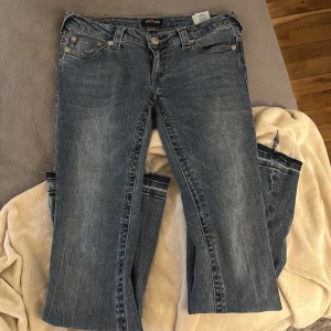 True Religion blå jeans, stl 28 - Säljer ett par klassiska blå jeans från True Religion i storlek 28. Jeansen har ikoniska bakfickor med broderad logga, femficksmodell och råa, fransiga benslut. Snygg tvättad denim med lätt slitning och raka ben. Perfekta för en cool och avslappnad look.