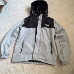 Grå och svart vindjacka från The North Face - Snygg grå och svart vindjacka från The North Face med huva och dragkedja framtill. Jackan har två fickor med dragkedja och justerbar huva. Perfekt för blåsiga dagar och enkel att matcha med streetwear. Funkar för M också! 