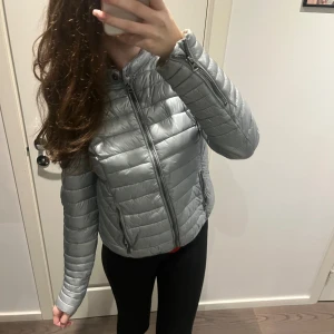 Silvrig pufferjacka från Uhip - Snygg silvrig pufferjacka från Uhip med quiltad design och hög krage. Jackan har dragkedja framtill, två fickor med dragkedja och ett emblem på ärmen. Perfekt för kyliga dagar och ger en clean look.