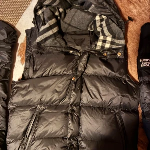 Svart dunväst med rutigt foder - Snygg svart dunväst med rutigt grått och svart foder i huvan. Västen har dragkedja och knappar framtill samt justerbar huva. Perfekt för lager-på-lager och chill höst- eller vinterstil.