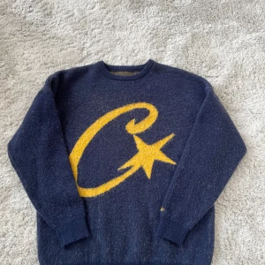 Mörkblå stickad tröja med gul stjärna från Corteiz - Säljer en mörkblå stickad tröja från Corteiz i storlek L. Tröjan har ett stort gult stjärnmotiv på framsidan och matchande gul detalj på ärmen. Materialet är 65% core yarn och 35% akryl, vilket gör den mjuk och skön. Rund hals och långa ärmar.