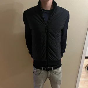 Marinblå J.Lindberg cardigan - Marinblå cardigan jacka från J.Lindeberg med quiltad front och stickade ärmar. Jackan har hög krage, dragkedja hela vägen och en snygg patch på ärmen. Perfekt för dig som gillar stilren design och vill ha något som funkar till många outfits.