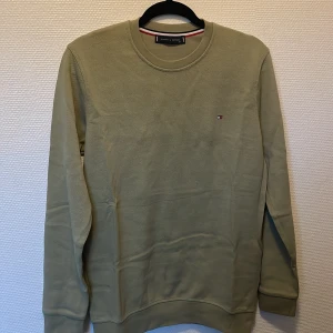 Olivgrön sweatshirt Tommy Hilfiger - Snygg olivgrön sweatshirt från Tommy Hilfiger i storlek S. Tröjan har rund hals, långärmade ärmar och ett litet broderat Hilfiger-logo på bröstet. Tillverkad i mjuk bomull med ribbade muddar vid ärmslut och nederkant. Perfekt för chill och streetwear.