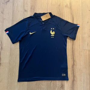 Frankrike Nike fotbollströja marinblå - Snygg marinblå fotbollströja från Nike med Franska landslagets emblem och två stjärnor broderade i guld på bröstet. Tröjan har korta ärmar, krage med knapp och diskret mönster i tyget. Materialet är lätt och andas, perfekt för match eller träning.