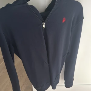 Marinblå hoodie från US Polo Assn - Säljer en marinblå hoodie från US Polo Assn med röd broderad logga på bröstet. Tröjan har huva med snörning, praktisk ficka framtill och dragkedja. Perfekt för chill dagar och har en skön, normal passform. Materialet är mjuk bomull.