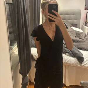 Svart omlottklänning från Cubus - Svart kort omlottklänning från Cubus i mjukt bomullsmaterial. Klänningen har v-ringad hals, korta ärmar med lätt volang och knyt i midjan för en snygg siluett. Perfekt för dig som gillar enkel och stilren design.