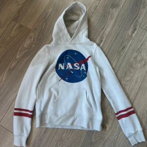 Vit NASA hoodie med röda detaljer  - Vit hoodie med stort blått NASA-tryck på bröstet och röda ränder på ärmarna. Klassisk känguruficka framtill och huva. Bekväm och snygg! 