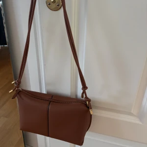 Brun axelväska med gulddetaljer från Zara - Nyskick! Brun axelväska i skinn med guldfärgade detaljer och dragkedja. Väskan har justerbara axelrem med knutar. Insidan är ljus och har en ficka med dragkedja. 