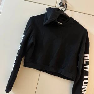 Svart croppad hoodie med tryck - Svart croppad hoodie med huva och vit text 'NEW YORK' längs ena ärmen. Tröjan har en avslappnad passform och är tillverkad i mjuk bomullsmix. Perfekt för en streetstyle-look och passar till både jeans och leggings.