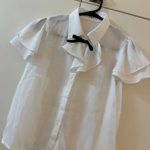 Vit volangskjorta med rosett från SHEIN - Supersöt vit skjorta från SHEIN med korta volangärmar, klassisk krage och svart rosett vid halsen. Skjortan har knäppning framtill och är gjord i ett lätt, tunt material som känns luftigt och fräscht.