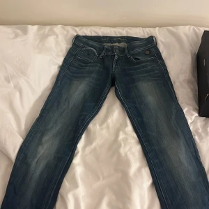 G-Star Raw 3301 blå jeans med dragkedja - Snygga blå jeans från G-Star Raw, modell 3301. De har klassisk femficksdesign, coola slitningar och en unik dragkedjedetalj på ena bakfickan. Märkespatch baktill och tydliga kontrastsömmar. Perfekt passform och stilren look.försetäller storlek xs