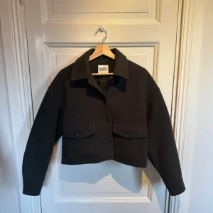 Svart croppad overshirt från Zara - Säljer en grön croppad overshirt från Zara med klassisk krage och stora fickor framtill. Jackan har knäppning med knappar och långa ärmar. Perfekt för dig som gillar en stilren och enkel look.