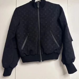 Svart bomberjacka Louis Vuitton - Svart bomberjacka från Louis Vuitton med ikoniskt monogrammönster ton-i-ton. Jackan har ribbade muddar, hög krage och två snedställda fickor med dragkedja. Materialet är mjukt och jackan har en snygg, modern passform. Perfekt för dig som vill sticka ut med lyxig streetstyle.