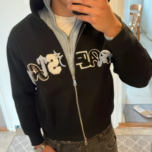 Svart Trapstar hoodie med broderi - Svart hoodie från Trapstar med stor broderad logga i blått, vitt och svart över bröstet. Tröjan har huva, dragkedja framtill och två fickor. Materialet är mjuk bomull och passformen är normal. Perfekt för dig som gillar streetwear och vill sticka ut.