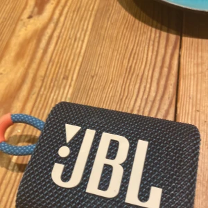 JBL Go 3 - Kompakt och portabel JBL Go 3 Bluetooth-högtalare i blått och orange. Snygg design med slitstarkt tyg och praktisk bärögla. Perfekt för resor och utomhusbruk. Ser ut att vara i mycket gott skick utan synliga skador eller slitage.
