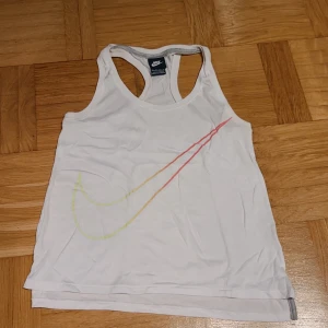Vit linnetopp från Nike XS - Säljer en vit linnetopp från Nike i storlek XS. Toppen har brottarrygg och ett stort Nike Swoosh-tryck i gradient från gult till rött på framsidan. Perfekt för träning eller sportiga outfits. Tillverkad i mjuk bomull för skön känsla.