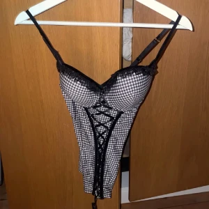 Rutig korsett med snörning och spets - Säljer en svartvit rutig korsett med smala axelband, dekorativ snörning framtill och spetsdetaljer vid bysten. Snygg passform och perfekt för att skapa en edgy look.