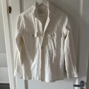 Vit overshirt från Jack & Jones - Stilren vit overshirt från Jack & Jones i bomull. Skjortan har klassisk krage, två bröstfickor med lock och knappar framtill. Perfekt att slänga över en t-shirt för en avslappnad look.