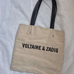 Canvas tote dag Voltaire & Zadig - Zadig & Voltaire canvas tote bag Stilren beige tygväska med svart tryck "VOLTAIRE & ZADIG" och svarta handtag. Perfekt för vardag eller shopping - lätt, trendig och rymmer mycket. ○ Bra skick överlag, men handtagen är något slitna och har sprickor (se bild),. Inga fläckar eller hål på själva väskan. 2 Pris: 450 ( Öppet för rimligt bud) Kan skickas eller hämtas i Stockholm/Solna