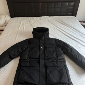 Svart dunjacka från Sail Racing XL - Säljer en svart dunjacka från Sail Racing i storlek XL. Jackan har en lång, boxig passform med stora fickor framtill, hög krage och huva. Materialet är slitstarkt och jackan är fodrad med dun för extra värme. Perfekt för kalla vinterdagar. Den är helt ny utan prislapp har även kvittot till den, den har aldrig använts