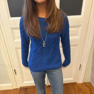 Blå stickad tröja med kabelmönster - Snygg blå stickad tröja med kabelstickat mönster och rund hals. Tröjan har långa ärmar och ribbade muddar vid ärmslut och nederkant. Perfekt för dig som gillar en klassisk men ändå trendig look.