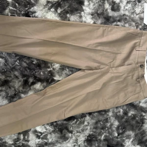 Beige kostymbyxor från Selected Homme - Snygga beige kostymbyxor från Selected Homme med rak passform och klassisk design. Byxorna har bälteshällor, sidfickor och stängs med knapp och dragkedja. Perfekta för en clean och stilren look.