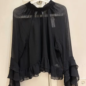Ny  kollektion Svart blus med volang och knyt - Supersnygg svart blus i transparent polyester med volangdetaljer längs nederkant och ärmslut. Blusen har lång ärm och en hög krage med knyt i nacken. Perfekt för dig som gillar en flowy och chic look med extra detaljer. Köpte online , jag köpte för 399kr 