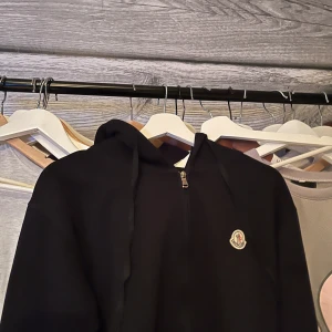 Moncler zip - Svart hoodie från Moncler med half zip och klassisk logga på bröstet. Tröjan har huva med snörning och är tillverkad i mjuk bomull. Perfekt för dig som gillar stilrena och exklusiva märken. Snygg passform och långärmad design. Nästan helt ny, kan fixa bättre bilder vid intresse!