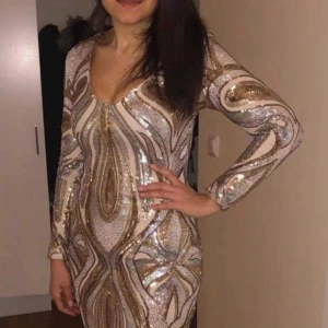 Glittrig guld och silver kortklänning - Säljer en superglittrig kortklänning med lång ärm och v-ringning. Klänningen har ett mönster i guld och silver paljetter på beige botten, vilket ger en riktigt lyxig och festlig vibe. Den är figurnära och har dragkedja i ryggen för enkel påklädning.