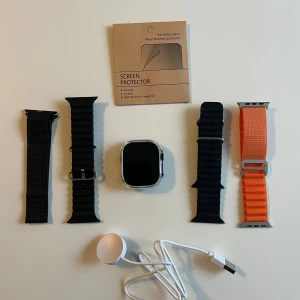 Apple Watch Ultra med extra armband - Apple Watch Ultra med fyrkantig silverfärgad boett och fyra utbytbara armband: svart gummiband med ribbad struktur, svart textilband och orange textilband med metallspänne. Ingår laddare och skärmskydd. Perfekt för dig som gillar teknik och vill ha en snygg smartklocka.