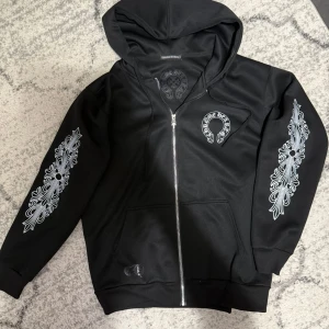 Svart hoodiejacka från Chrome Hearts - Svart hoodiejacka från Chrome Hearts med dragkedja och ikoniska silvriga tryck på ärmarna. Jackan har huva, två fickor fram och detaljerad metallzipper med Chrome Hearts-logga. Materialet är mjukt och bekvämt, perfekt för en streetwear-look.