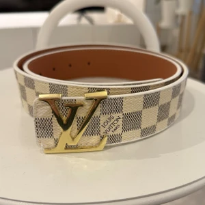 Rutig skinnbälte med ett schyst spänne - Säljer ett snyggt bälte i skinn med klassiskt rutigt mönster i beige och grått. Bältet har ett stort guldfärgat spänne och detaljerad präglad logga. Perfekt accessoar för att lyfta din outfit med exklusiv känsla.