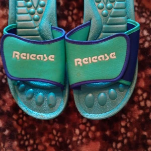 Turkosa sandaler från Release - Säljer ett par turkosa sandaler från Release med breda remmar och vit logga. Sulan har mönstrad struktur för extra komfort och grepp. Remmarna är i syntetmaterial med blå och gröna detaljer. Perfekt för chill dagar och strandhäng.