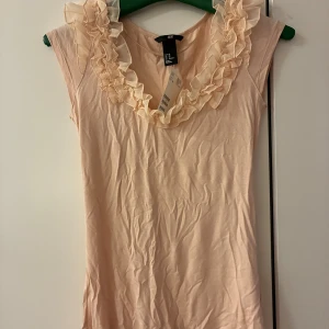 Ljusrosa/aprikos blus med volang från H&M - Supersöt ljusrosa blus från H&M i storlek XS. Toppen har volangdetaljer runt halsringningen och är ärmlös. Materialet är mjukt med 95% viskos och 5% elastan, medan volangerna är i 100% polyester. Perfekt för dig som gillar romantiska och feminina detaljer. Tunn, lätt och stretchig! Prislapp kvar