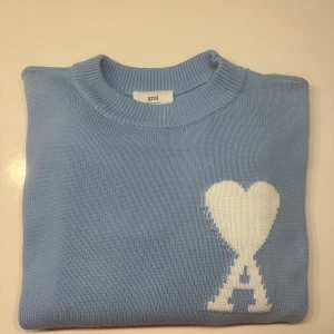 Ljusblå stickad tröja från Ami - Snygg ljusblå stickad tröja från Ami med rund hals och korta ärmar. På bröstet finns ett stort vitt hjärta och bokstaven A som ger en unik vibe. Tröjan har ribbade muddar och är perfekt för dig som gillar stilrena detaljer.