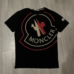 Svart Moncler t-shirt med stor logga - Svart t-shirt från Moncler med stor, ikonisk logga i vitt och rött framtill, dekorerad med små paljetter. Klassisk rund hals och korta ärmar. Moncler-märke på ena ärmen. Snygg och trendig design som sticker ut.