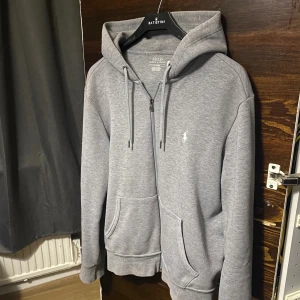 Grå hoodie från Polo Ralph Lauren - Säljer en stilren grå hoodie från Polo Ralph Lauren med dragkedja, huva och snörning. Tröjan har två stora fickor framtill och den klassiska broderade loggan på bröstet. Perfekt för en chill och avslappnad look.