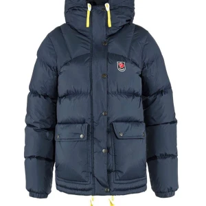 Mörkblå dunjacka från Fjällräven - Snygg svart dunjacka från Fjällräven med huva, gula snören och stora fickor framtill. Jackan har klassisk pufferstil, tryckknappar och dragkedja. Perfekt för kalla dagar och riktigt skön att bära. Kan skicka fler bilder. Aldrig använd ligger kvar i förpackning samt skick 10/10